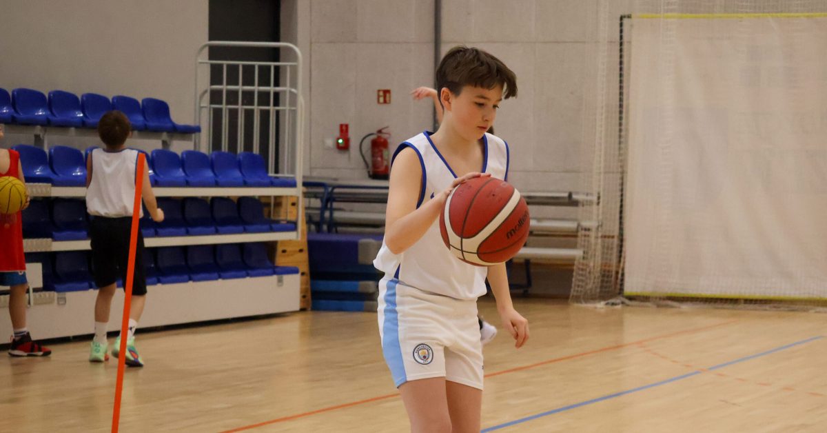 Chłopiec w białym stroju koszykarskim z niebieskimi detalami kozłuje piłkę na hali sportowej. W tle puste trybuny i ściana hali.