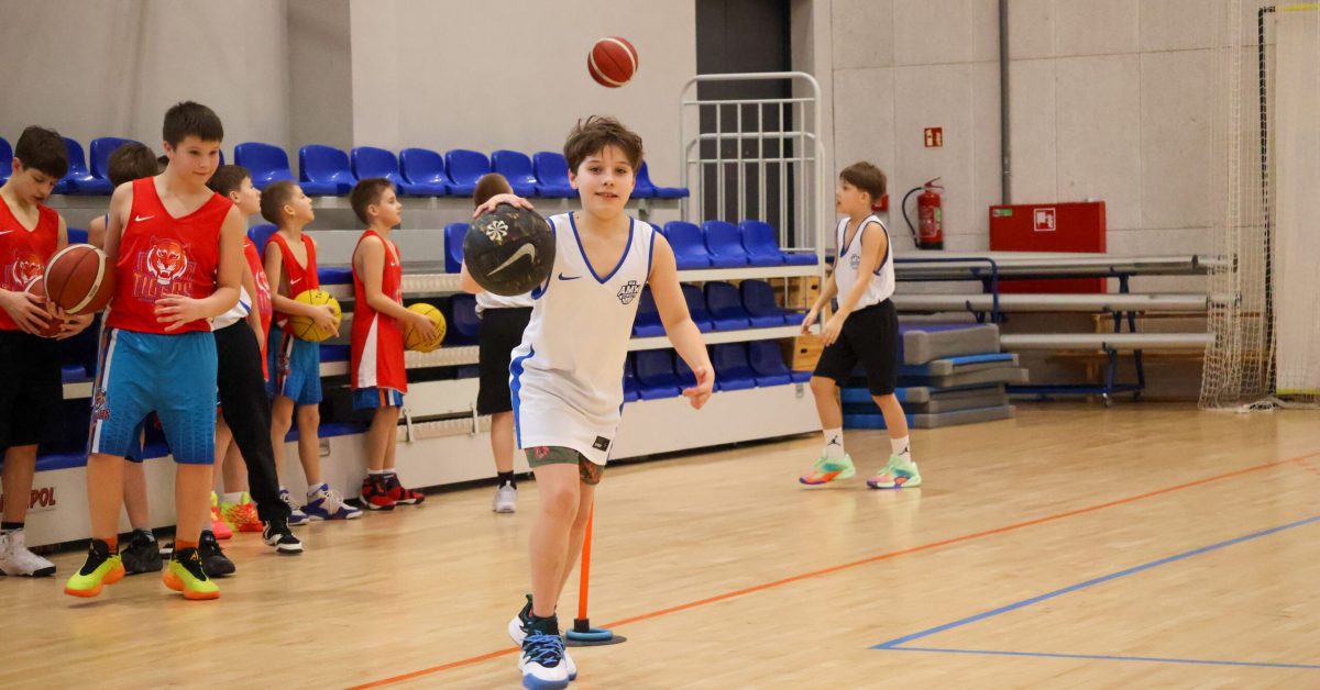 Chłopiec w białym stroju z niebieskimi elementami wykonuje kozłowanie z piłką do koszykówki. W tle inni zawodnicy czekają na swoją kolej.