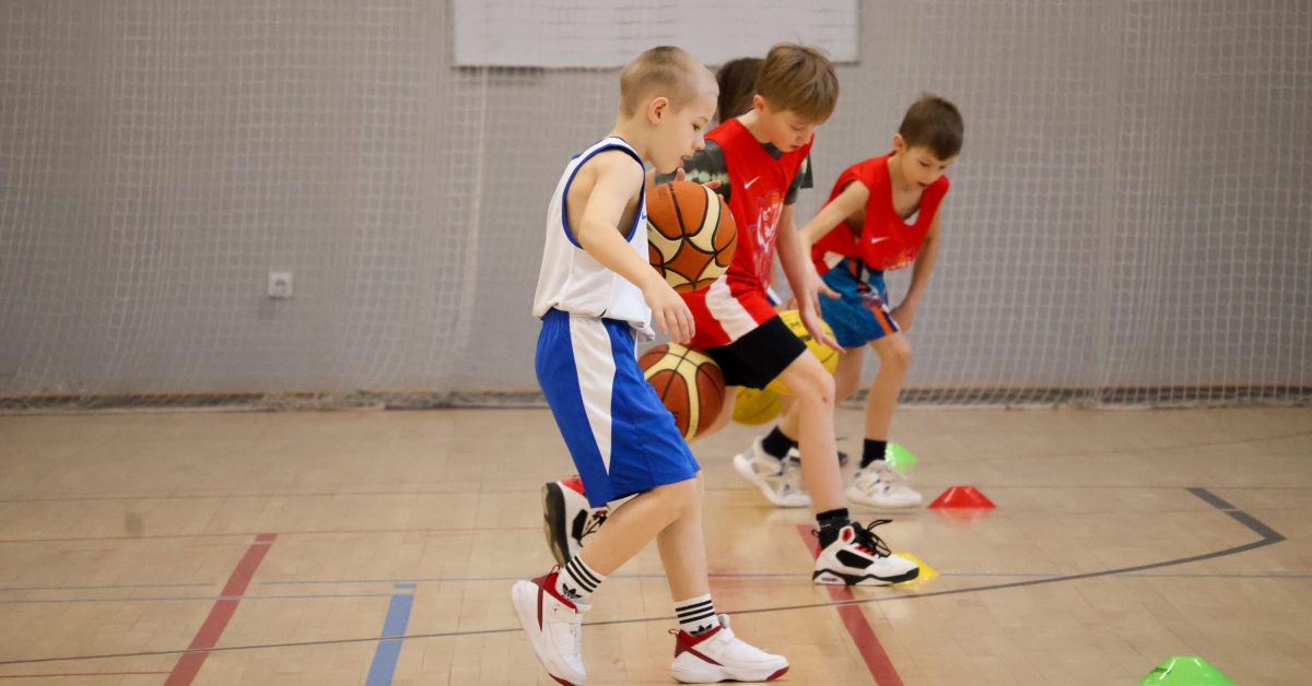 Kilku chłopców, ubranych w białe i czerwone stroje, biegnie po boisku, omijając rozstawione pachołki podczas ćwiczeń treningowych z piłką koszykarską.