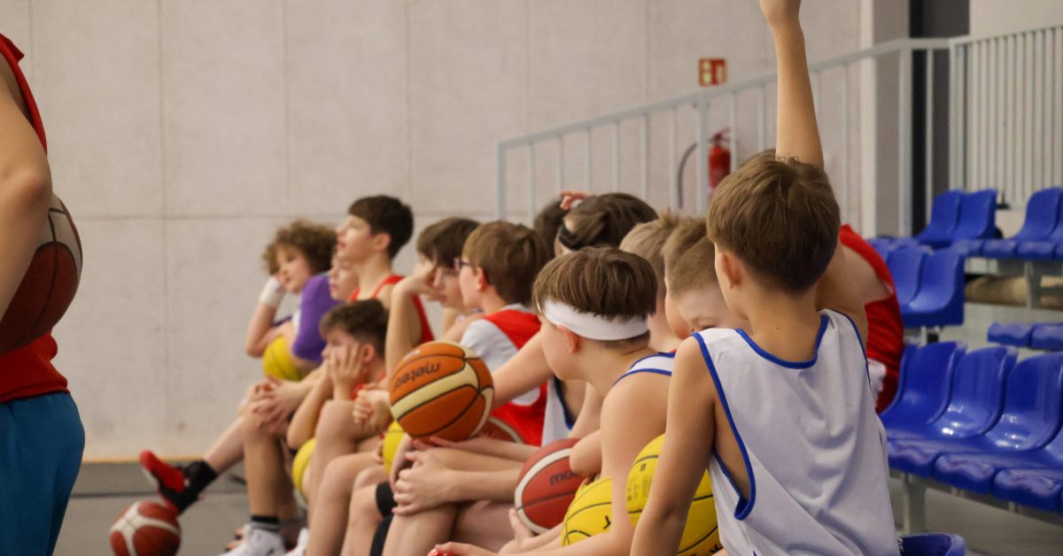 Grupa młodych koszykarzy siedzi na ławce podczas treningu, jeden z nich podnosi rękę. Wszyscy uważnie obserwują trenera.