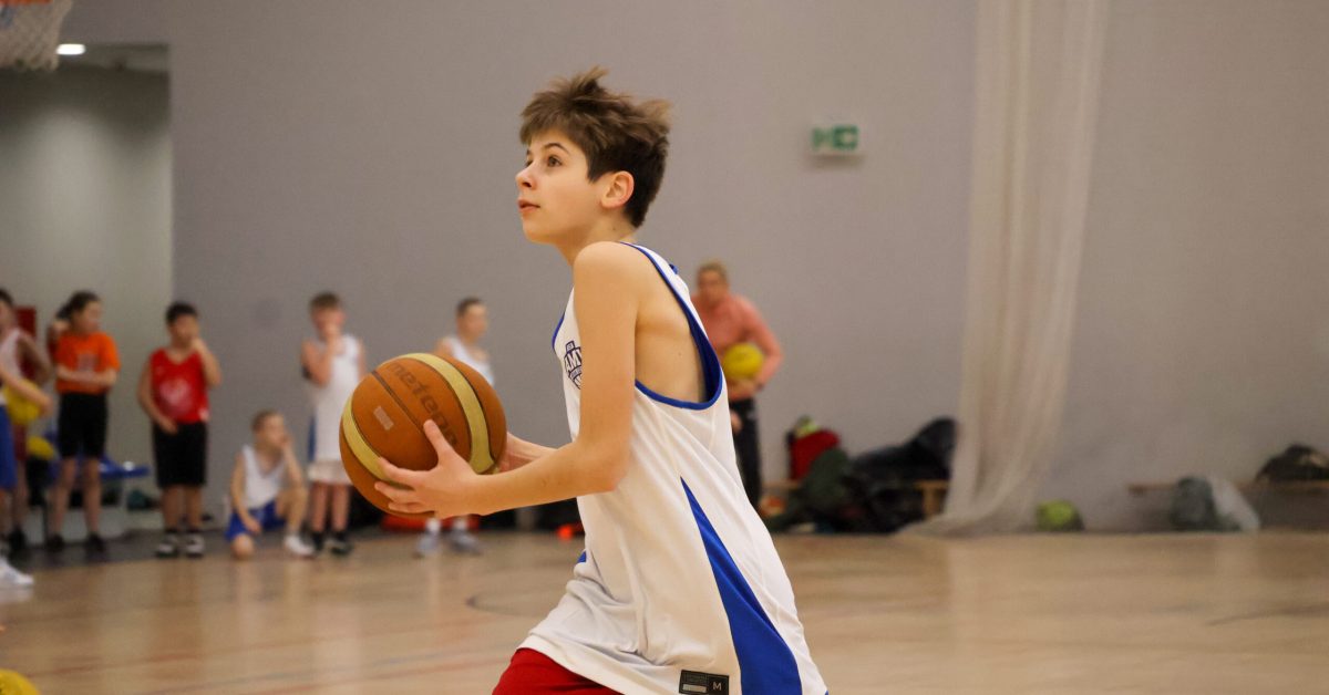 Młody koszykarz w białym stroju z czerwonymi elementami biegnie z piłką po hali sportowej, przygotowując się do rzutu. W tle widać innych zawodników.