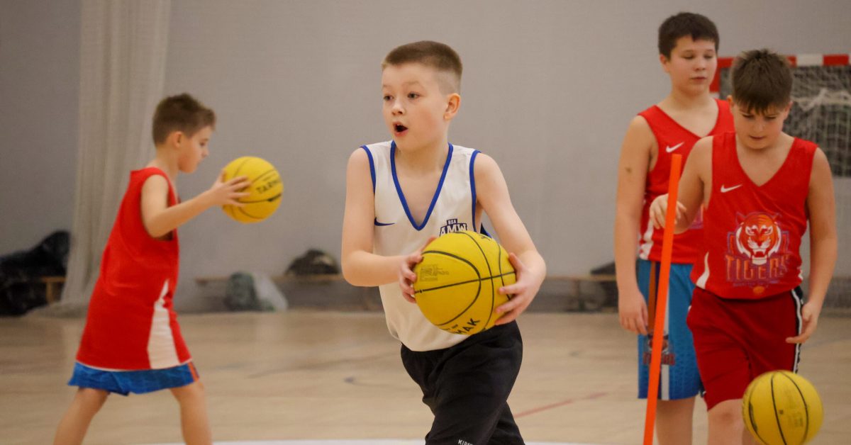 Chłopiec w białym stroju z czarnymi spodenkami trzyma żółtą piłkę i przygotowuje się do rzutu, podczas gdy inni zawodnicy ćwiczą w tle.