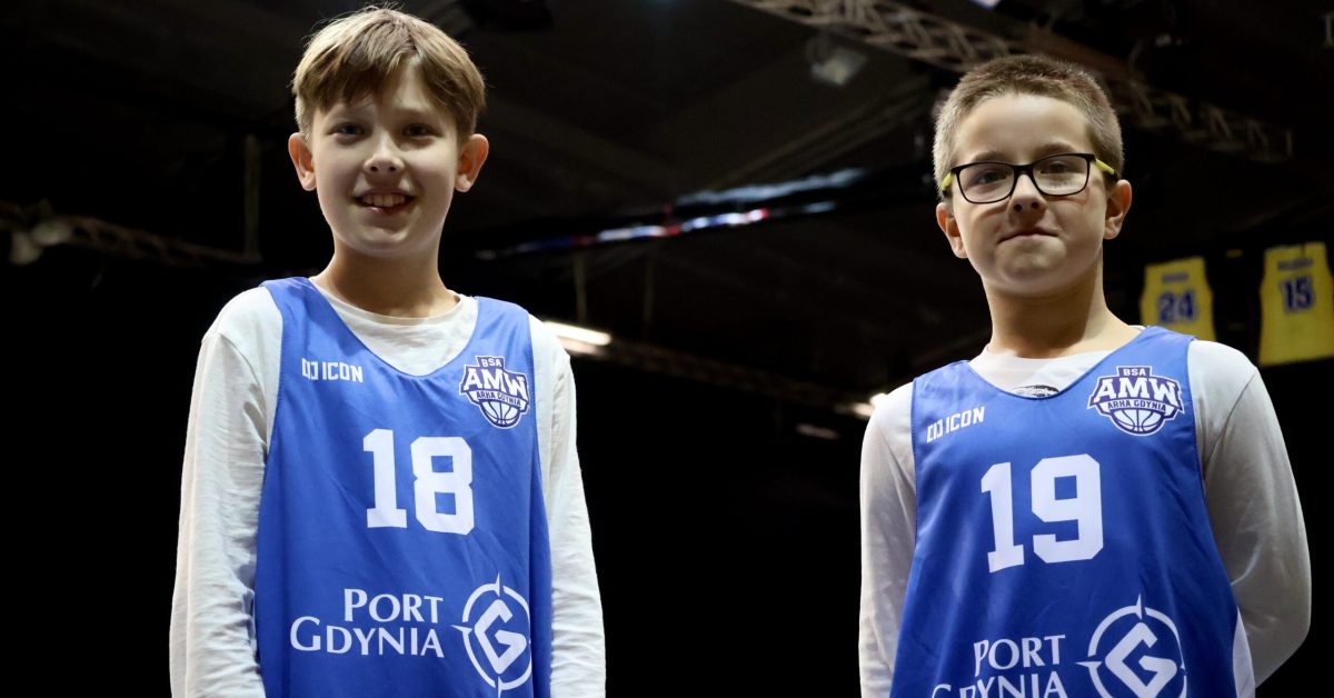 Dwóch młodych chłopców w niebieskich koszulkach z numerami 18 i 19 pozuje do zdjęcia, uśmiechając się, na tle hali sportowej.