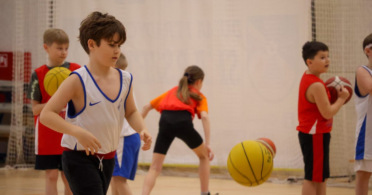 Młody zawodnik w białym stroju kozłuje piłkę, a wokół niego ćwiczą inni dzieci ubrani w czerwone i pomarańczowe stroje sportowe.