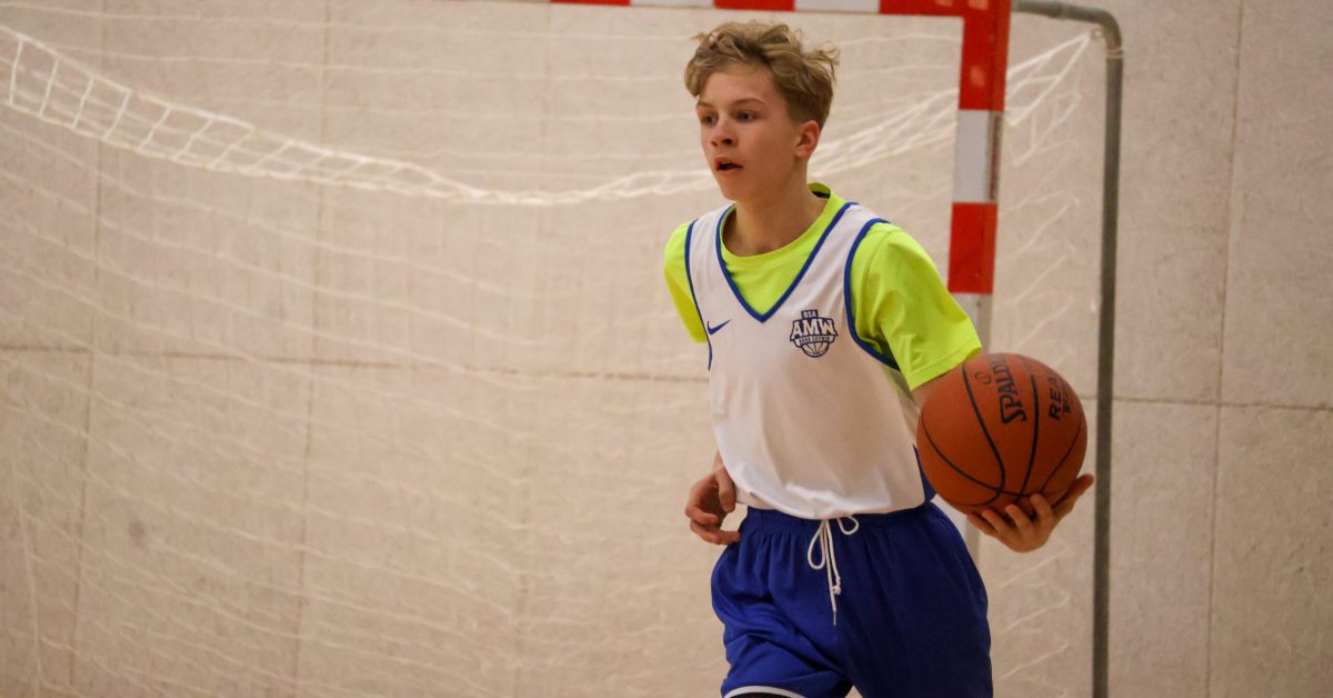 Chłopiec w białym stroju z zielonymi elementami trzyma piłkę do koszykówki i biegnie na hali.