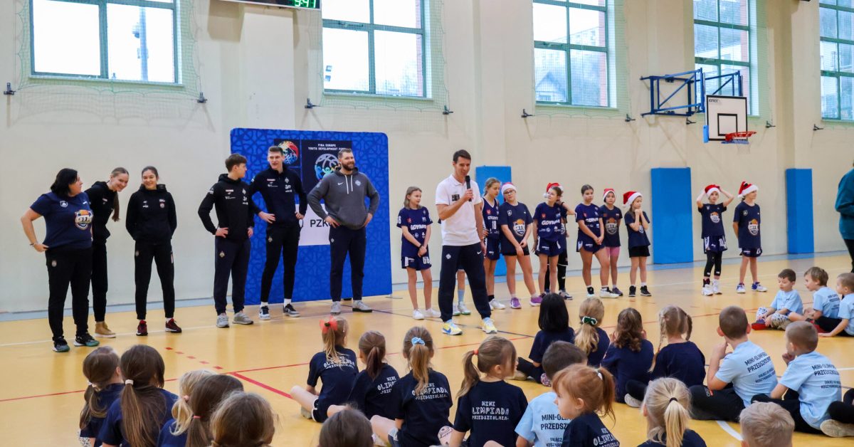 Trener w czarnym stroju przemawia do grupy dzieci siedzących na podłodze sali gimnastycznej, za nim baner i inni trenerzy.