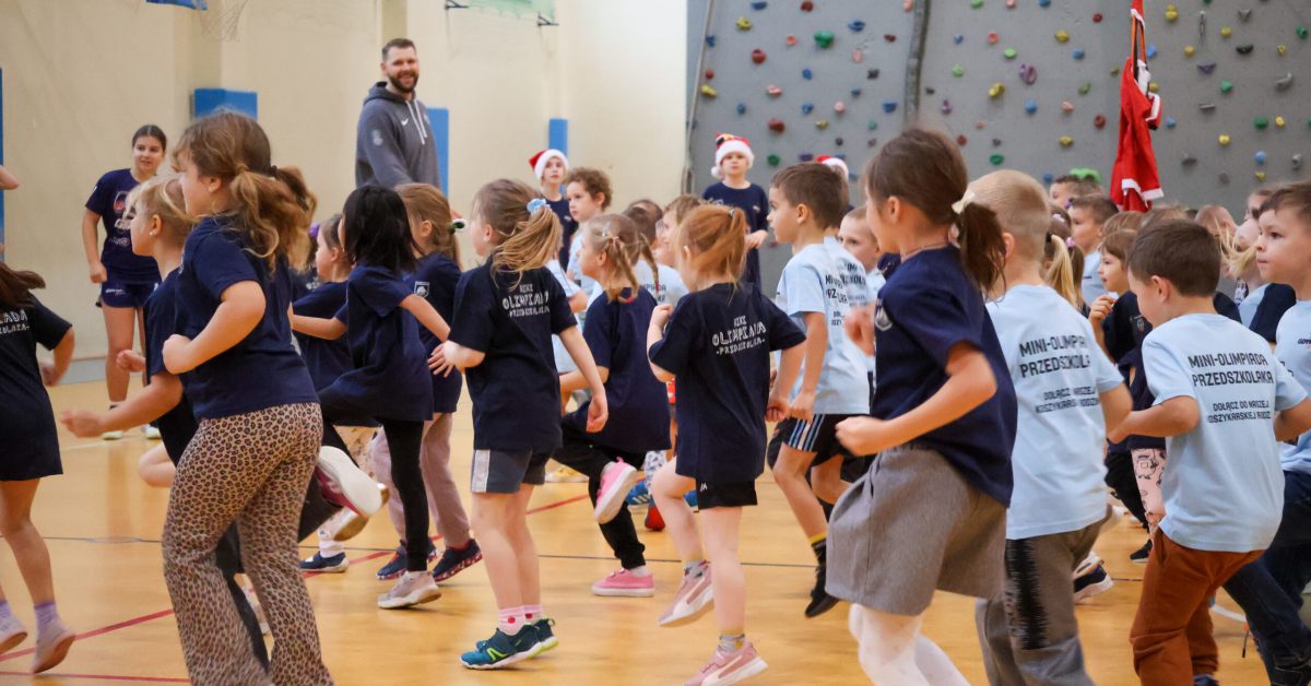 Grupa dzieci energicznie wykonuje ćwiczenia ruchowe pod okiem trenerów na hali sportowej.