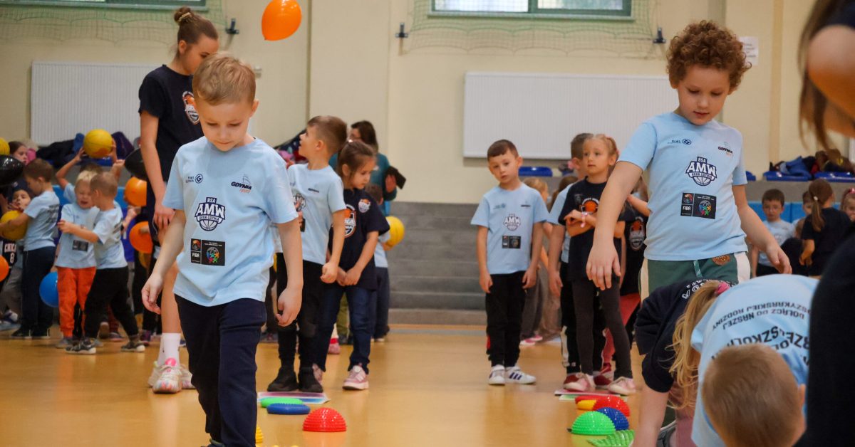 Dzieci w koszulkach sportowych biorą udział w zabawie ruchowej na sali gimnastycznej, wśród kolorowych przeszkód i piłek.
