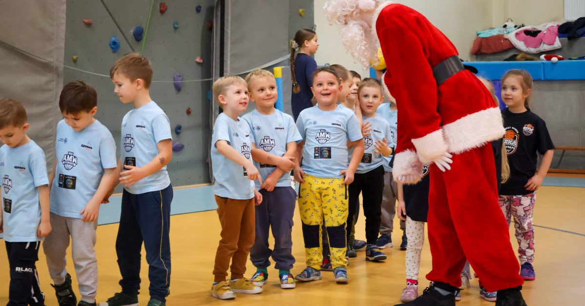 Święty Mikołaj pochyla się, rozmawiając z grupą dzieci ubranych w jasnoniebieskie koszulki sportowe.