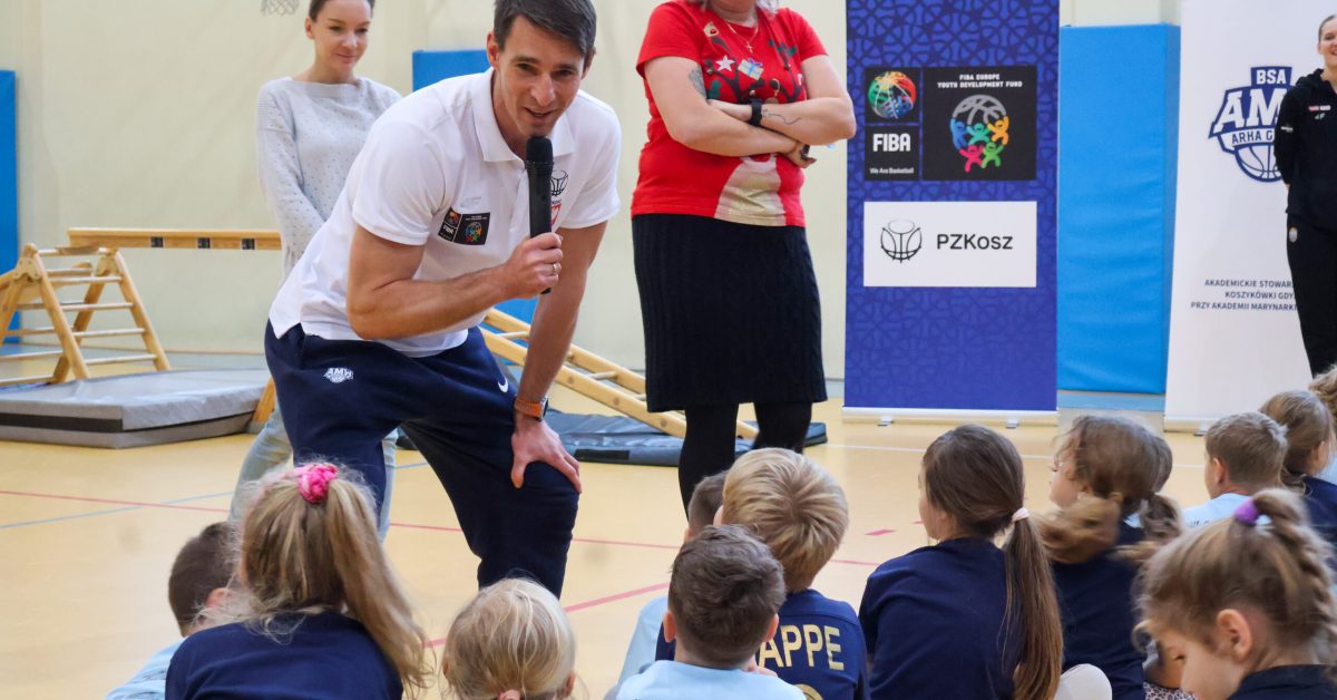 Trener klęczy przed grupą dzieci, rozmawiając przez mikrofon, podczas gdy nauczycielka obserwuje sytuację w tle.